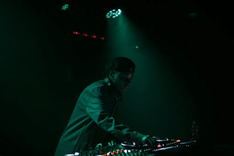 A DJ performs behind a mixing console under green stage lighting.